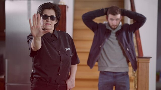 Serious confident police officer making stop gesture standing in emergency lights indoors with sad bandit at background holding hands behind head. Law offence consequences and robbery prevention.