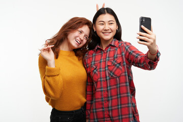 Two women friends. Making selfie on smartphone. Girl plays with hair and put horns to a friend. Wearing yellow sweater and checkered shirt. People concept. Stand isolated over white background