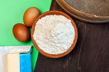 a wooden rolling pin, two eggs, wooden bowl filled with flour, sieve, stick of butter and black cutting board on green background