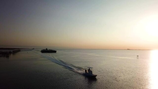 Aerial drone view of small flat bottomed fishing boat speeding ahead of large green barge that is follow the Gulf Intercoastal Waterway thru Laguna Madre to Port Corpus Christi; Texas; at sunset