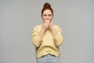 Portrait of frowning, adult redhead girl with hair gathered in a bun. Wearing pastel yellow sweater and jeans. Cover her mouth with palms. Watching at the camera isolated over grey background
