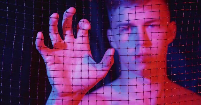 Isolation anxiety. Life challenge. Comfort zone barrier. Defocused portrait of disturbed doubtful worried man creasing mesh fence with hand palm in pink blue neon light on dark background.