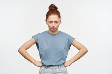 Portrait of upset, adult redhead girl with hair gathered in a bun. Wearing blue t-shirt and jeans. Put hands on her hips and frowning. Watching at the camera isolated over white background
