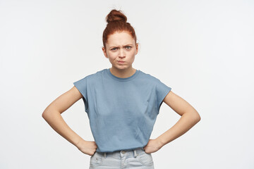 Fototapeta premium Serious looking woman, girl with ginger hair gathered in a bun. Wearing blue t-shirt and jeans. Put hand on her hips and frowns. Emotion concept. Watching at the camera isolated over white background
