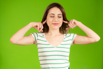 Young beautiful woman wearing casual t-shirt standing over isolated green background covering ears with fingers with annoyed expression. Deaf concept.