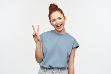 Portrait of cute, adult redhead girl with hair gathered in a bun. Wearing blue t-shirt and jeans. Showing peace sign, tilted her head and smile. Watching at the camera isolated over white background