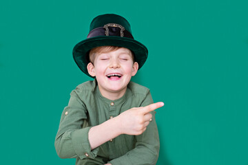 A closeup portrait of smiling preteen boy squeezed eyes shut in carnival St. Patrick's Day hat pointing finger text space on green backgroundwhite. Copy space.