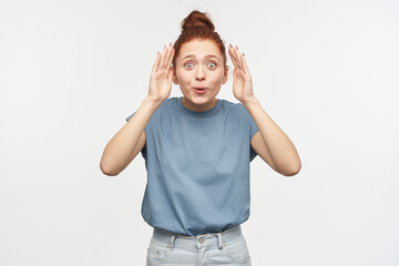 Nice looking woman, playful girl with ginger hair gathered in a bun. Wearing blue t-shirt and jeans. Playing hide and seek, uncover her face. Watching at the camera isolated over white background