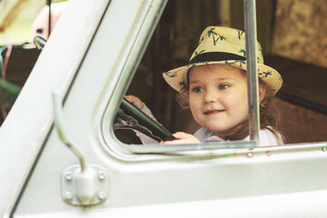 Little girl in a hat sits behind the wheel of an old disassembled car