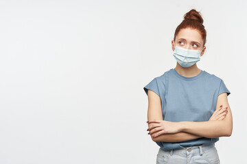 Teenage, wonder looking woman with ginger hair gathered in a bun. Wearing blue t-shirt and protective face mask. Watching with crossed hands to the left at copy space, isolated over white background