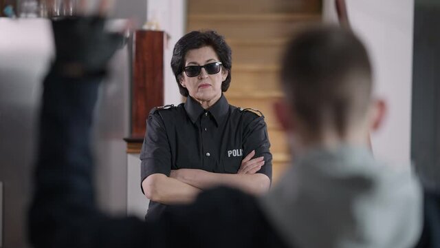 Confident policewoman in sunglasses crossing hands looking at criminal with hands up. Portrait of Caucasian female police officer arresting burglar indoors. Confidence and law protection.