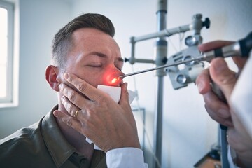 Patient at ENT clinic. Specialist doctor - Otolaryngologist doing nose examination of young man. - obrazy, fototapety, plakaty