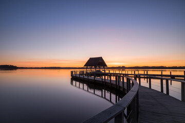 Swinging curved Pier construction and shelter with thatched roof at beautiful colorful sunrise under clear sky at Lake Hemmelsdorf, Schleswig-Holstein, Northern Germany