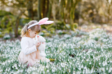 Adorable little girl with Easter bunny ears holding soft plush toy in spring forest on sunny day,...