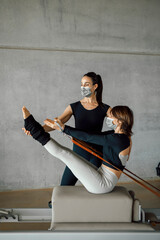 Woman in protective face mask taking Personal Pilates lesson on a Reformer machine, doing v-sit crunches, at a big fitness center, wide industrial gym studio. Healthy fitness lifestyle.
