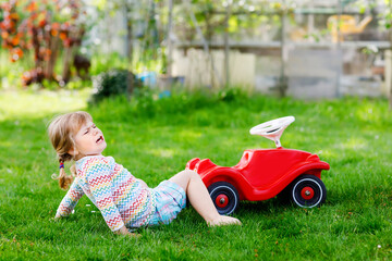 Little active adorable toddler girl falling down from toy car, having pain and crying, outdoors. Upset child on summer day. Injured hurt kid in gaden