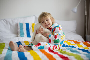 Happy toddler child, blond boy with colorful bathrobe, sitting in bed with stuffed toy after bath, smiling