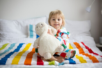 Happy toddler child, blond boy with colorful bathrobe, sitting in bed with stuffed toy after bath, smiling