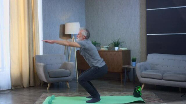 Portrait of a gray-haired senior man squatting on a yoga mat. Home fitness exercise squats on his feet. Grandfather in excellent athletic body shape. Old man in sportswear. Health in old age.
