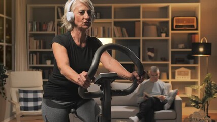 active mature elderly woman exercise bike workout at home,caucasian old female in headphones workout on bicycle listen to music in living room apartment night time shot
