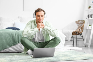 Handsome young man with laptop and headphones meditating at home - obrazy, fototapety, plakaty