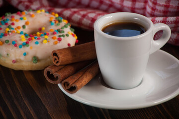 A cup of aromatic black coffee and coffee beans on the table with cinnamon sticks. Morning espresso coffee for breakfast in a beautiful white cup. With a delicious donut in the background.