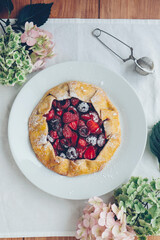 Delicious freshly baked vegan strawberry and cherry galette on wooden rustic background with hydrangea flowers, top view. Sweet food, summer dessert.