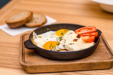 Hearty breakfast is served in a black frying pan and wooden board, fried eggs with bacon and tomatoes, two pieces of bread are on a wooden table