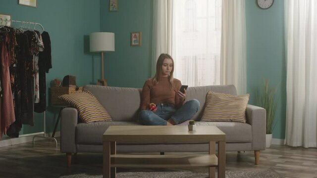 Lady in blue jeans and brown sweater sits on beige sofa on window background, holding red apple and looking at something on phone. On coffee table is glass jar with green apples. Dolly in.