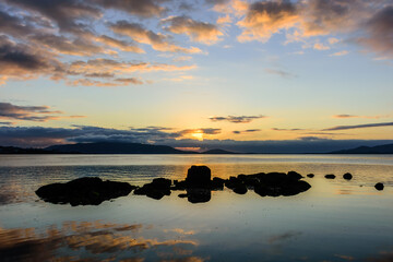 Seascape at sunrise on the horizon, calm sea and rocks in the foreground, blue sky and golden clouds - 419670432