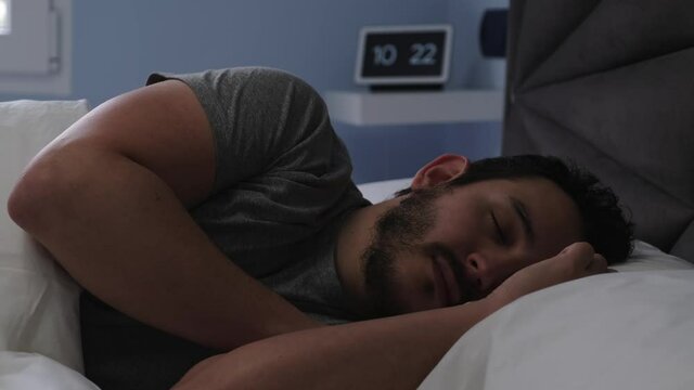 Young latin man sleeping in his bed late in the morning. Digital clock at the background. 4k video.