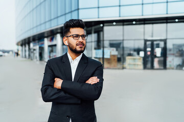 Fototapeta premium A modern office worker, an Indian man, a bank employee in a black suit, behind a large wooden building