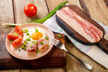 Fried eggs with bacon with tomato and green onion and on a wooden table in a plate next to chopped bacon and tomatoes.