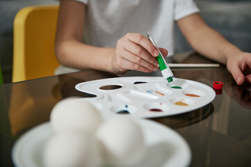 Little boy's hand squeezing green paint from a tube onto a palette, blurry eggs on a saucer in the foreground. Closeup.