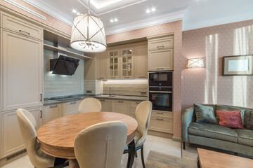 Spacious kitchen with the large lounge area with a soft velvet olive sofa. Neoclassic beige kitchen with built-in appliances. The round  table with soft chairs standing in the center of the kitchen