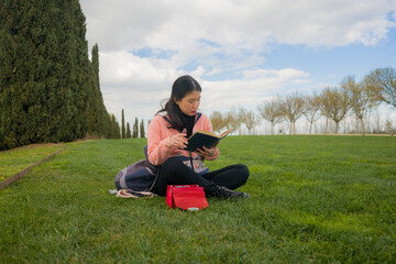 Fototapeta premium young Asian woman enjoying novel on grass - lifestyle portrait of young happy and pretty Chinese girl reading a book at beautiful city park in reading and studying concept
