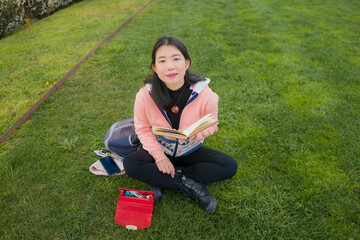 young Asian woman enjoying novel on grass - lifestyle portrait of young happy and pretty Japanese girl reading a book at beautiful city park in reading and studying concept