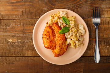 Baked chicken breast in sauce with salad on a plate next to a fork on a wooden table.
