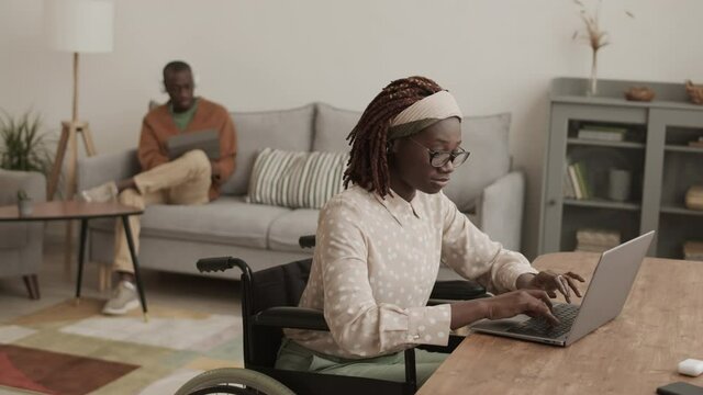Lockdown of young African disabled woman sitting on wheelchair at desk in living room, working on laptop and then looking at camera