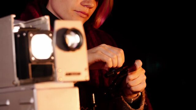 Close-up of an old fashioned slide projector and a young woman inserting its supplements for making ready to work. The caucasian lady is loading the photo negatives into a retro cinematographic device