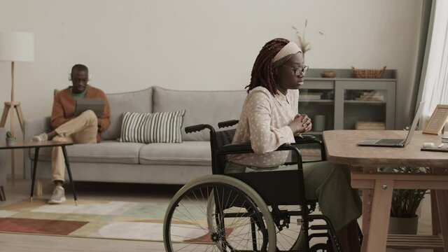 Lockdown of young African disabled woman sitting on wheelchair at desk in living room and having online business meeting using laptop