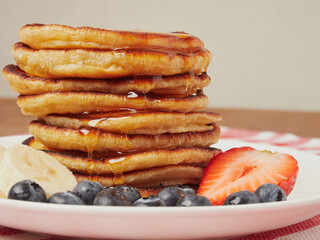 Close-up of delicious pancakes, with fresh blueberries, strawberries and maple syrup on a light background. With space to copy. Sweet maple syrup flows from a stack of pancakes