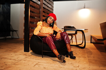 Fototapeta na wymiar Elegant afro american woman in red french beret, big gold neck chain polka dot blouse and leather pants sit on pouf.