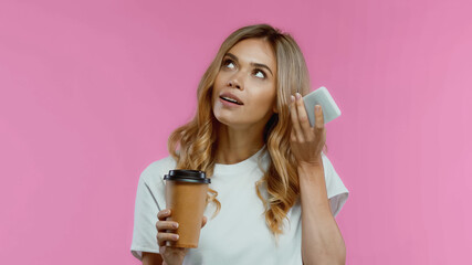 curious woman using smartphone while listening voice message and holding coffee to go isolated on pink