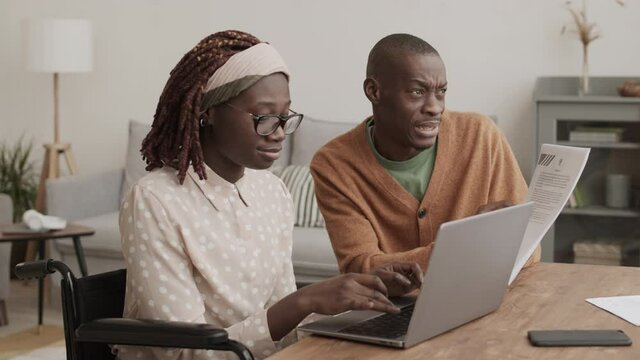 Medium shot of young African man sitting at desk with document in hand while young African disabled woman sitting on wheelchair and typing on laptop keyboard