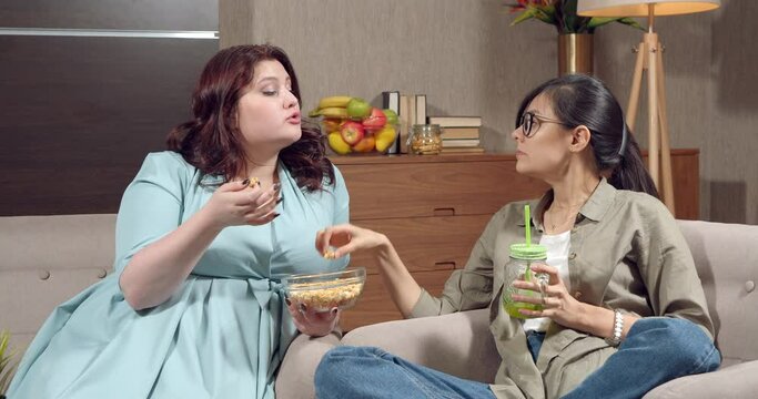 Two women friends eating popcorn while watching tv and discussing a movie sitting in the living room at home