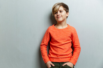A smiling boy 9 years old with a fashionable hairstyle in an orange sweater looks to the side. Grey background. Space for text.