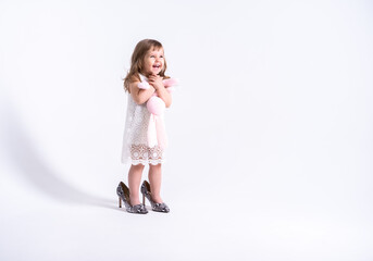 Funny little girl in white dress trying her mother shoes with heels on white background