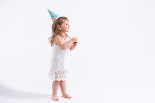 Funny child girl in white dress and birthday hat blowing in whistle on white background