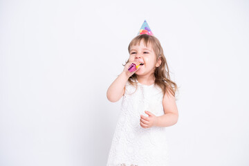 Funny child girl in white dress and birthday hat blowing in whistle on white background